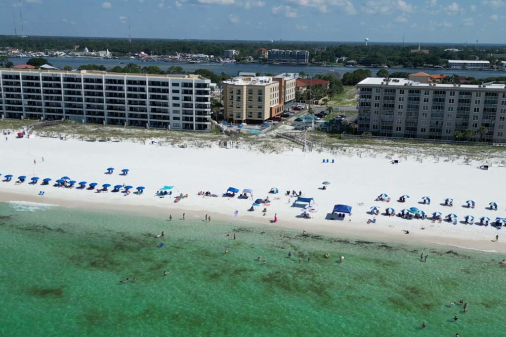 Aerial view of a beach hotel with white sand, green water, umbrellas, and seaside buildings.