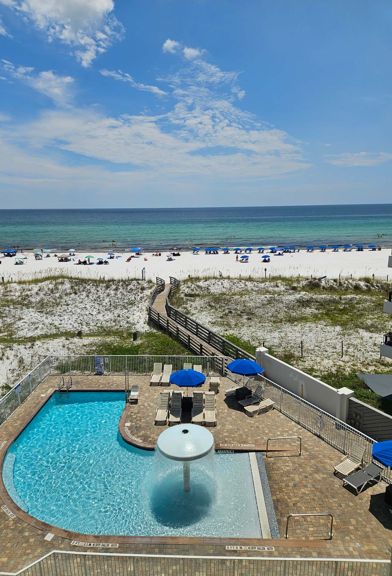 A pool with blue umbrellas at Beach Hotel overlooks a sandy beach and ocean under a cloudy sky.