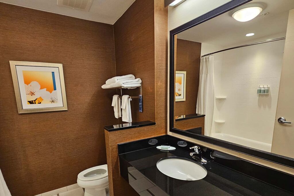 Modern hotel bathroom with brown walls, mirror, sink, toilet, towels, and bathtub with shower curtain.