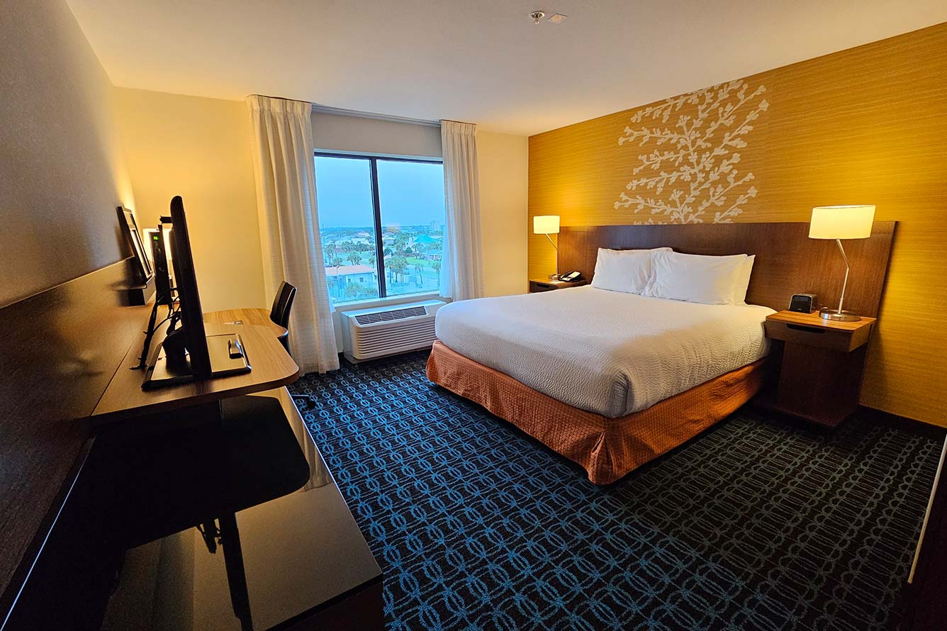 A Wavecrest Lodge hotel room with a large bed, desk, TV, lamps, and a sunlit window.