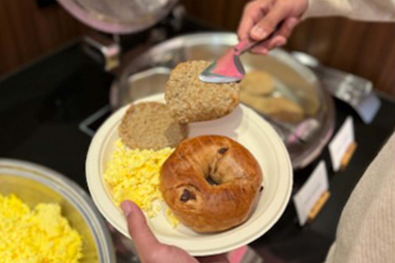A hand holds a plate with scrambled eggs and bagel at the Wavecrest Lodge buffet.