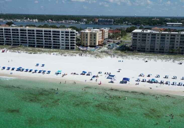 Aerial view of a beach hotel with white sand, green water, umbrellas, and seaside buildings.